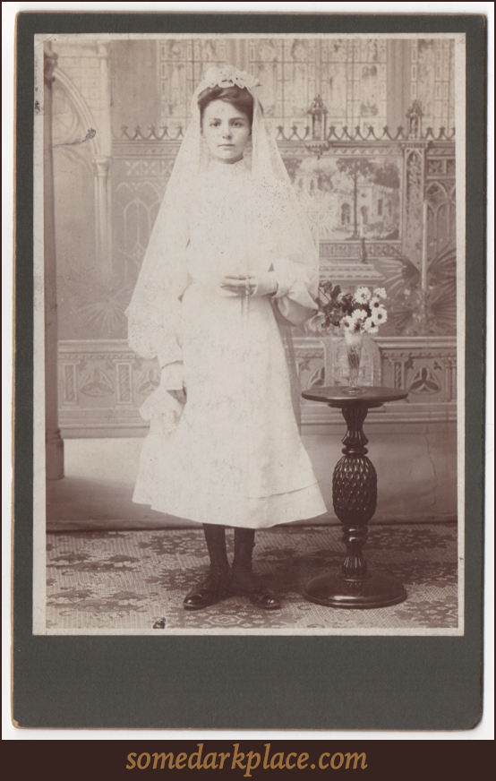 A young woman in a veil and wearing a white dress.  Most likely this is a Confirmation dress. She is holding a rosary.