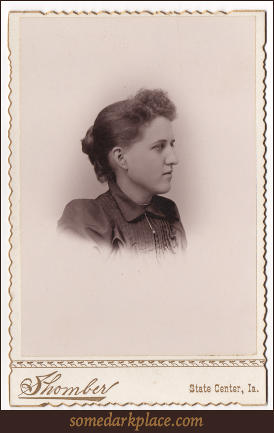 A bust image of a young woman in a dark dress or blouse with a small dangling pendant. Her hair is done in a bun at the back of the head and is loose curls in the front.