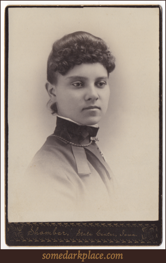 An attractive young woman in a high collared smock or dress. She is wearing a choker style necklace on the outside of the collar with a metal clasp at her throat. She is also wearing a broach on her left side.