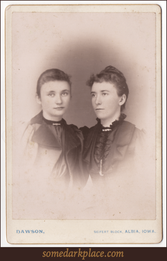 Two young women. One somewhat older that the other. Both are in partial profile. Both are wearing formal clothes with one in an overcoat and the other in a dress. Both have broaches at their necks.