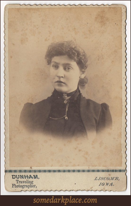 An attractive curly haired young woman with her hair pulled back. She wears both a ribbon around her throat with buttons or stones as well as an elaborate pendant with chains. She is wearing dark clothes and a sort of jacket.
