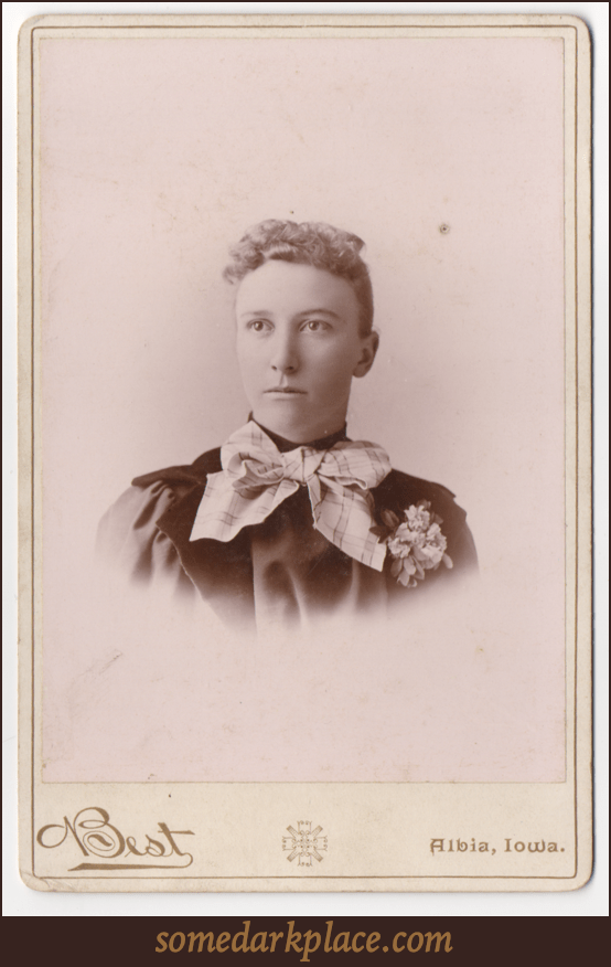 A young woman with her hair pulled back and curls in the front. She is wearing a a giant striped bow around her neck and a corsage over her heart. She is wearing a dark dress. This is a bust shot.