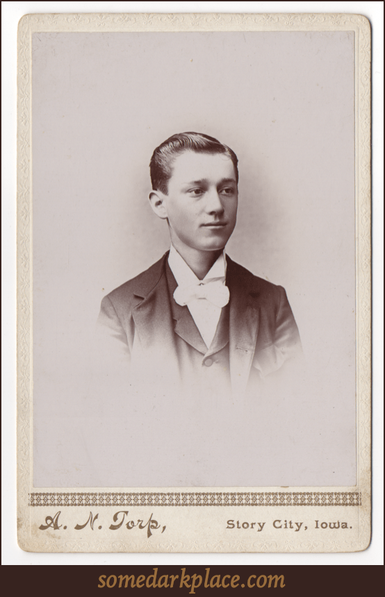 A dapper young man in formal wear. He has a dark suit and a white shirt with a large bow tie. His hair is short, parted on the left, and he has no facial hair.