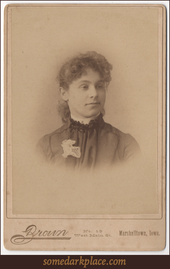 A curly haired woman with her hair swept back. She is wearing a pleated dress with a white lace flower on her lapel. She has a stiff white collar. 