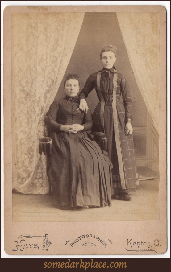 Two women. One seated, the other standing with her arm resting on the other's shoulder. Both wear full length dresses with one being a simpler dark dress with buttons down the front. The other with pleats and a pattern.