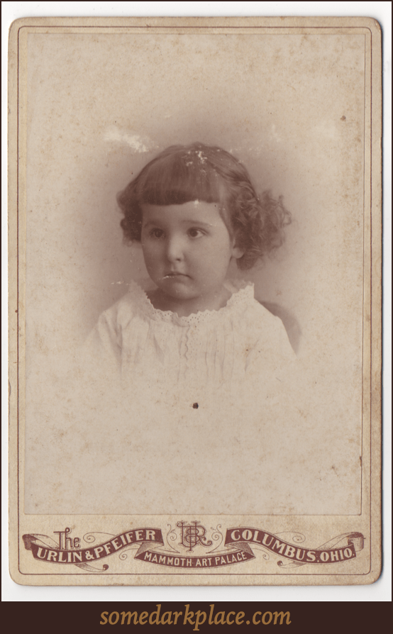 A toddler in a lacy dress or over shirt. She has short curly hair and unevenly trimmed bangs. She appears to be sitting.