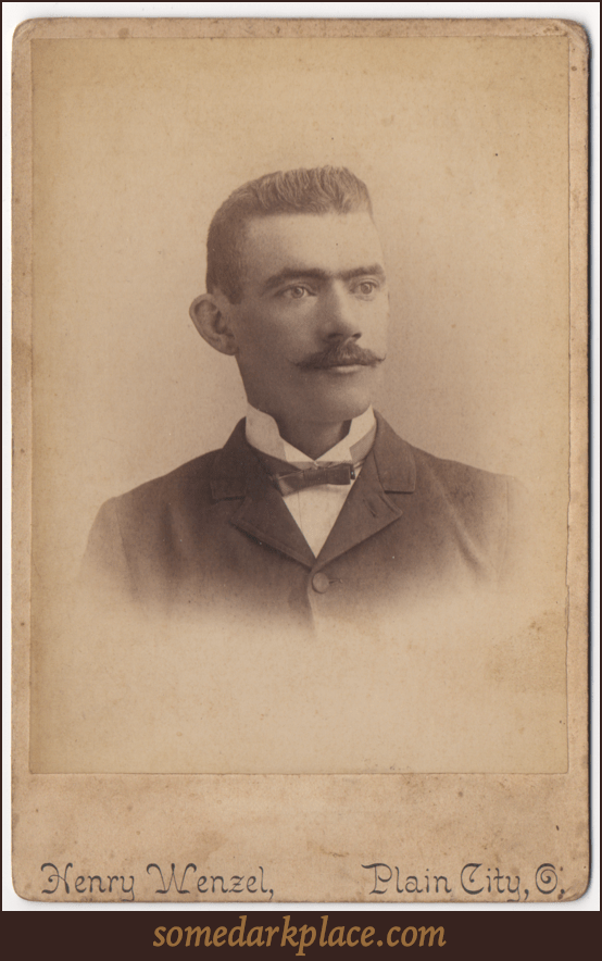 A dapper gentleman in a bowtie and a dark suit. His undershirt is light colored and his color is upturned. His hair is well groomed and short in a buzzcut style. He has a mustache that is turned up at the ends.