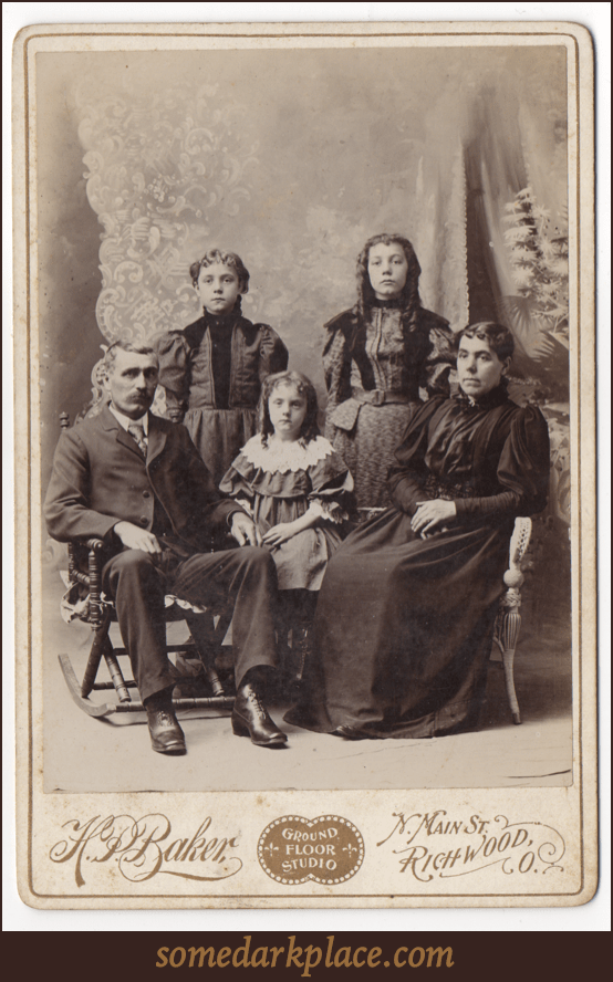 A family of five with a man, woman, and three children. All are dressed up wearing formal clothes. The adults are seated.