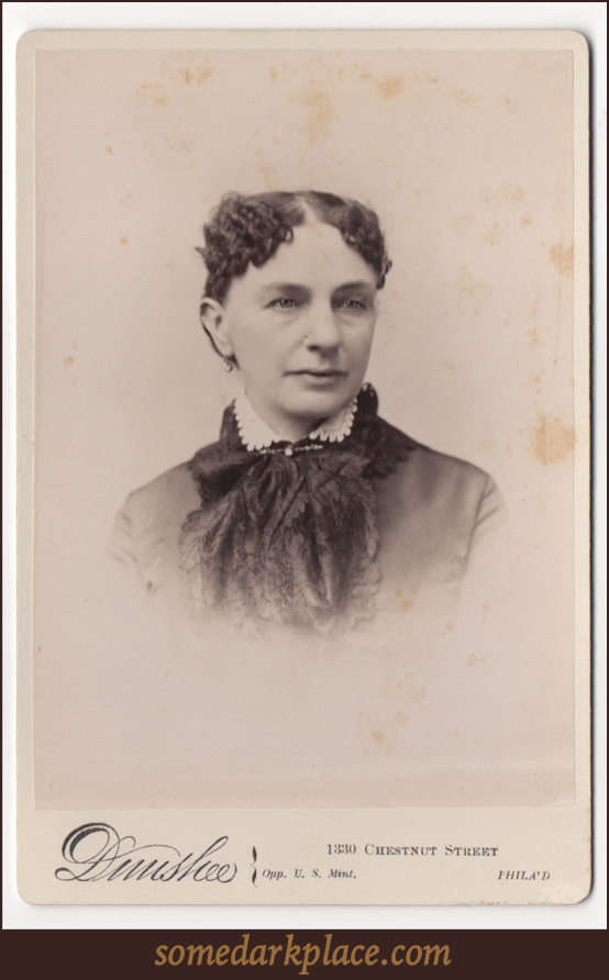 A woman with dark curly hair parted down the middle. She is wearing a pendant style earring. She is wearing a dress with a light colored scalloped collar. She has a clasp at her throat. She has a scarf or decorative cloth going down the front of her dress.