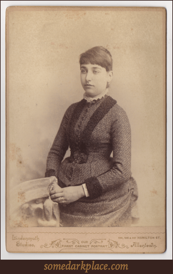 A young woman seated next to a prop stone pedestal. Her hands are filed before her. She is wearing a textured dress with a rounded and frilly collar. Her hair is pinned tightly against her head and her hair is pulled back. Her bangs are cut straight.