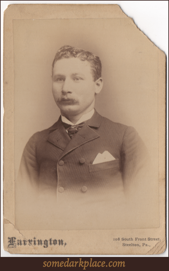 A dapper young man in a textured lined suit jacket and tie with some sort of pin through it. His hair is parted on the left and fairly short. He is looking slightly off camera. He has a mustache.