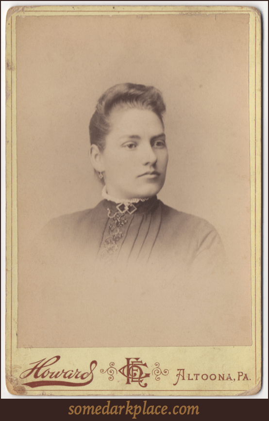 A young woman with an oval shaped face. Her hair it styled in a standing wave and is pulled back. She has a pendant earring and is wearing a rounded color trimmed in a ruffle or lace. She has a double diamond piece of jewelry as a clasp at her throat.