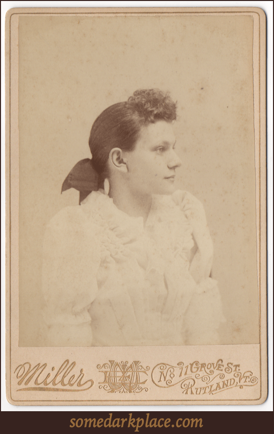 A profile of a young woman in an elaborate white dress with puffy sleeves, frills, and pleats. Her hair is curly in front and  pulled back into a large dark bow.