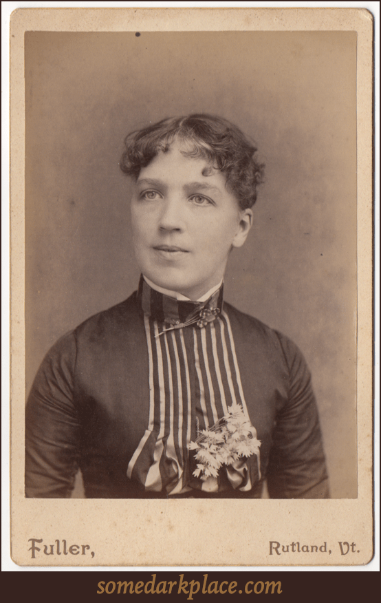 A woman with short dark curly hair. She is wearing a dress with pleats down the front and flowers on her chest. She has a high collar with a broach or clasp at her throat.