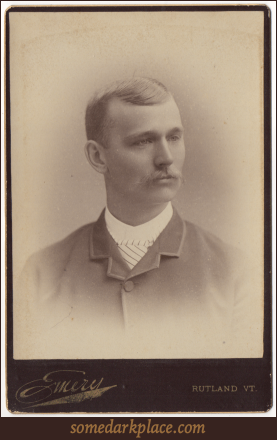 A well groomed young man with a light buttoned up suit coat and a striped tie. He is wearing a white shirt with a collar. His hair is parted on the left and is neatly trimmed. He sports a longer mustache.