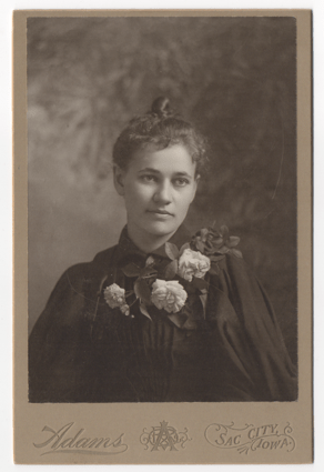 A beautiful young woman with her hair in a topknot and pulled back. She is wearing a dark dress and perhaps an overcoat or cloak. Her outfit is embellished with four large white roses and leaves.