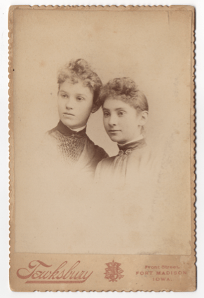 Two young women. Both in partial profile. Both are wearing dresses. One has a broach and the other a clasp at her throat. They wear high collars with white trimming.