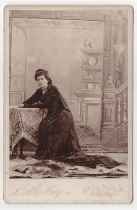 An obvious studio shot of a younger woman in dark flowing garb. She is wearing a headdress and has a cover around her throat. Her hair is curly, parted on the left, and pinned back. She sits at a prop table in front of a painted background.