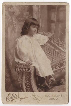 A young girl in an all white dress and black boots. She has long hair that is curly on the ends, but otherwise hangs straight down. She is seated in a whicker chair in a studio in front of a painted backdrop.