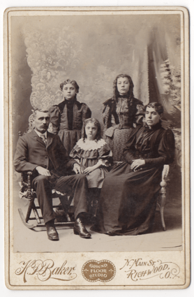 A family of five with a man, woman, and three children. All are dressed up wearing formal clothes. The adults are seated.