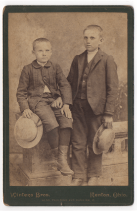 Two young boys sitting on a stone railing in front of a painted background in a studio shot. Both are holding stew hats with ribbons around the brim. One child is older than the other by a year or two.
