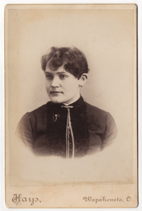 A young woman with short unkempt hair. She has a masculine appearance. She wears a top with buttons and piping down the front. She has a clasp at her throat and another pice of ornamental jewelry.