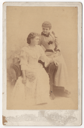 Two attractive young women. One fully seated and the other leaning into the armrest of this chair. One woman is looking at the other affectionately. Both women are wearing formal dresses.
