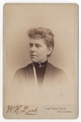 A woman with her hair pinned back and piled atop her head. Her bangs are curly and she looks like she would have a lot of hair to let down if she were so inclined. Her collar is rounded and dark and she has a piece of jewelry of some sort at her neckline.