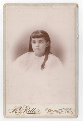 A young girl in a white robe or smock. Could be a sleeping shirt or more likely a dress. Her hair is long with some curl to it. Her bangs are neatly trimmed and even. Most her hair falls down her back, but one curled strand sits on her shoulder.