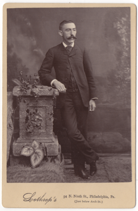 A man in a full dark suit leaning against a prop of a garden wall. He has on a light colored dress shirt and dark shoes with a shine to them. His mustache is large and impressive and well groomed.