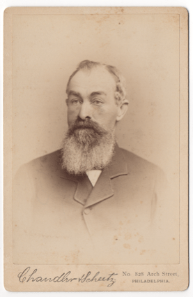 An older bearded gentleman wearing a suit coat and undershirt. He does not appear to be wearing a tie, but this could be obscured by his beard.
