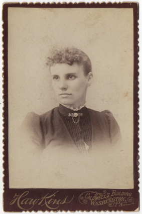 A curly haired young woman with a high collared dress with lace trim and a clasp and chain at her throat. Her dress is pleated in the front and has a piece of jewelry hanging in the front.