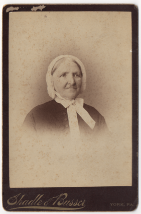 A old woman in a bonnet tied in a bow around her neck. Some sort of broach or clasp is at the center of the bow. Her gray hair is pulled back, parted down the middle, and mostly covered. She is wearing a dark frock with large buttons down the center.