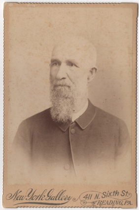 An older gentleman with white facial hair. The image has either faded or has exposure issues, so is difficult to tell if he is bald or just has shorter white hair. He is wearing a simple overcoat with a lighter shirt peaking out.