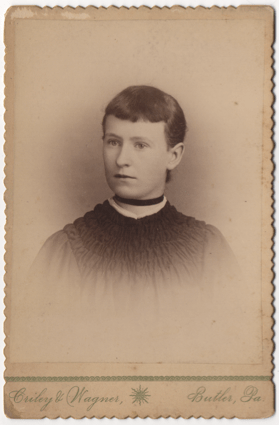 A short haired young woman with dark straight hair parted on the left. She is wearing a dress and does not have pierced ears. She wears a ribbon around her throat like a choker.