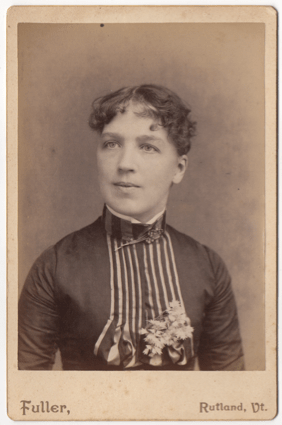 A woman with short dark curly hair. She is wearing a dress with pleats down the front and flowers on her chest. She has a high collar with a broach or clasp at her throat.
