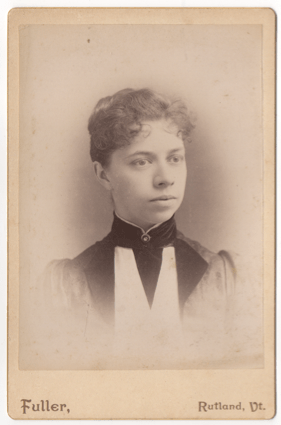 A woman with short or pulled back curly hair wearing a dress with a high collar and a clasp with a stone in the center. Her dress has large lapels.