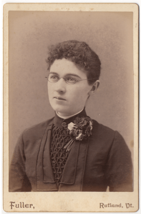 A young woman in a dark dress or top. The front has an intricate cloth lattice in a triangular pattern. He has a corsage and a clasp at the front of her collar. She is wearing glasses and has short curly hair or it is pulled back.