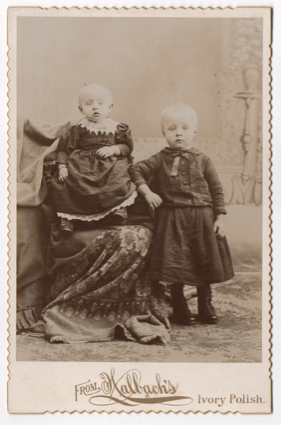 Two young tow headed children, both wearing dresses. Most likely two boys, but the infant could be either.