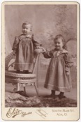 Two small children in matching dresses. One is standing in a chair, the other beside. The younger of the two appears to be a boy, the other a girl.…