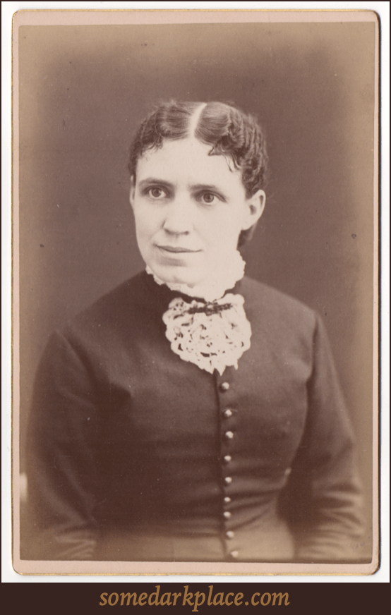 A woman with a severe part down the middle of her head with her hair pulled back tightly. She has a wide mouth and a wide nose. She is wearing a dark dress with shiny buttons down the front.