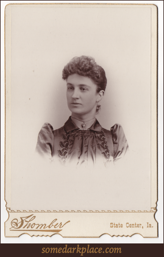 A young woman with her hair pulled back and curls in the front. She is wearing a choker necklace with a heart shaped center stone surrounded by smaller stones. 