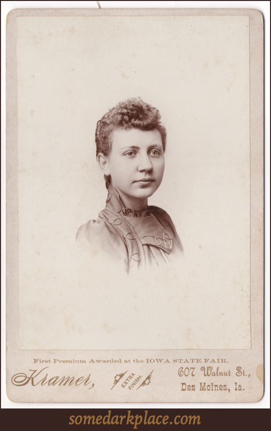 A headshot of an attractive young woman perhaps in her late teens. She is wearing a dress with a high collar and elaborate decorative piping. Her hair is curly in the front and pulled back or short.