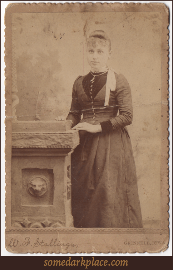 A young woman standing against a painted studio backdrop leaning on a prop pedestal with bas-relief carving. She appears to be about college age and is wearing a full length dress with buttons down the front.