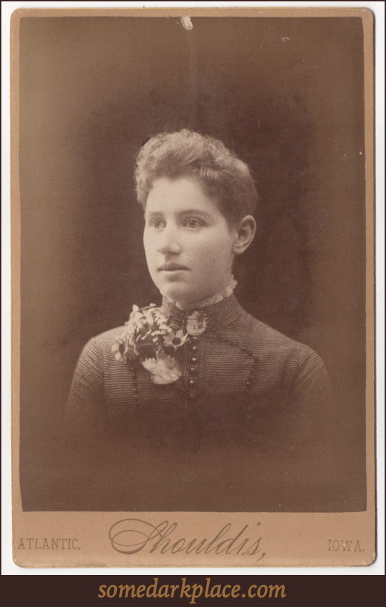 A young woman with short hair in high collared dress with a lace ruff. The dress is austere and almost military in appearance with large buttons and embellishments. Her face is unlined and her ears appear to be unpierced.