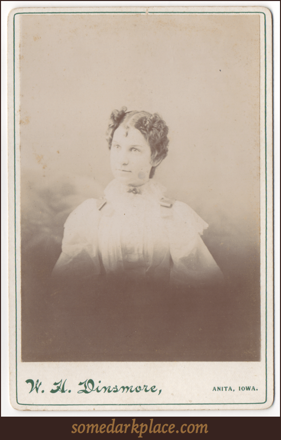 A young woman with a severe part down the middle of her head. She is wearing all white. She has decorative buckles or ribbons on her shoulders and a broach at her throat.