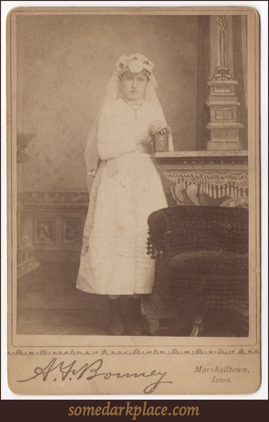 A young girl dress in a white first confession dress. These outfits have a similar appearance to wedding dresses. She is clutching a small prayer book or Bible.