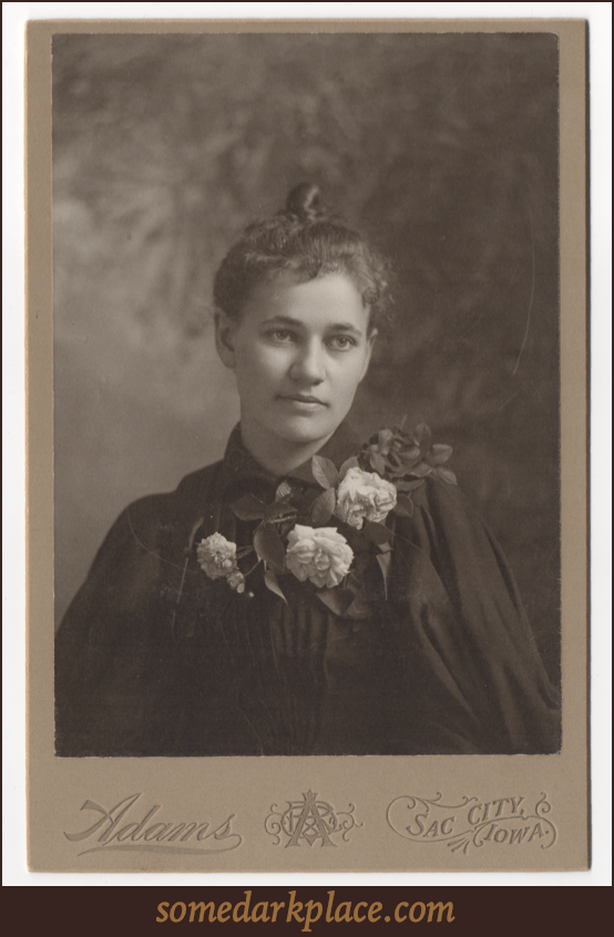 A beautiful young woman with her hair in a topknot and pulled back. She is wearing a dark dress and perhaps an overcoat or cloak. Her outfit is embellished with four large white roses and leaves.