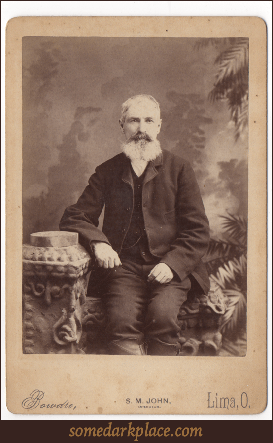 An older gentleman with impressive facial hair. He has gone mostly gray except for his mustache. He is wearing a dark suit and has a visible watch chain. He is seated.