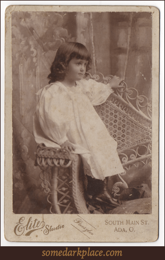 A young girl in an all white dress and black boots. She has long hair that is curly on the ends, but otherwise hangs straight down. She is seated in a whicker chair in a studio in front of a painted backdrop.